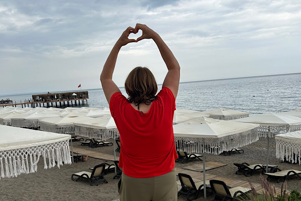 Anita steht am Meer unter bewölktem Himmel und formt mit erhobenen Armen ein Herz – Symbol für Herzöffnung, Mut und den Weg in die innere Freiheit.