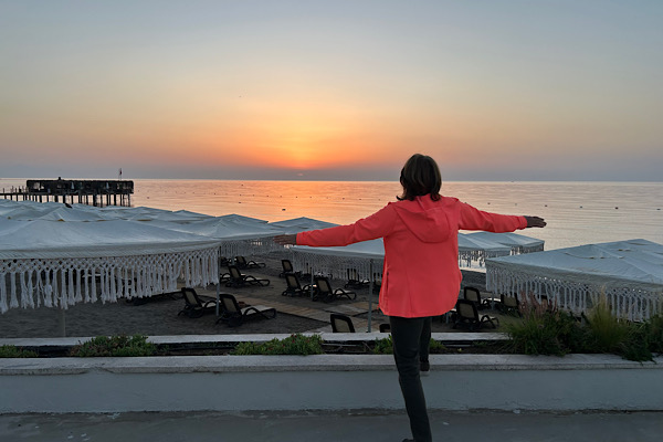 Anita steht am Meer im Sonnenaufgang und breitet die Arme weit aus – Symbol für Aufbruch, Inspiration und den Beginn eines neuen inneren Weges.