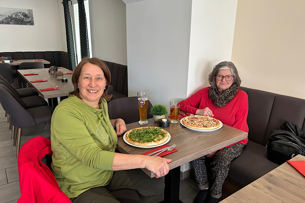 Karin und Anita sitzen am Tisch in der Pizzeria und genießen gemeinsam ihre Pizzas.