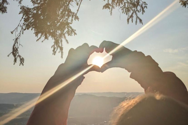 Hände formen ein Herz im Sonnenlicht, umgeben von Blättern und Berglandschaft – Symbol für emotionale Öffnung und innere Klarheit.