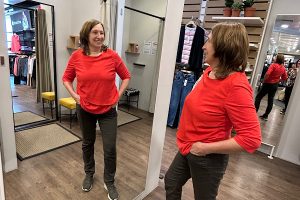 Anita Griebl steht vor einem Spiegel, von vorne und von der Seite sichtbar, im roten Shirt, nachdenklich und nicht in die Kamera blickend.