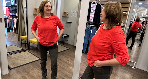 Anita Griebl steht vor einem Spiegel, von vorne und von der Seite sichtbar, im roten Shirt, nachdenklich und nicht in die Kamera blickend.