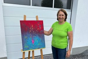 Anita Griebl steht im Garten neben ihrem intuitiv gemalten Energie‑Bild und lächelt ruhig – Ausdruck von Kreativität, Achtsamkeit und heilsamer Energiearbeit.