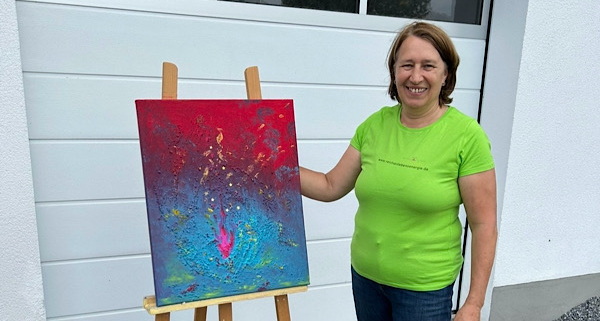 Anita Griebl steht im Garten neben ihrem intuitiv gemalten Energie‑Bild und lächelt ruhig – Ausdruck von Kreativität, Achtsamkeit und heilsamer Energiearbeit.
