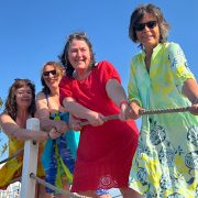 Vier Frauen in bunten Strandkleidern stehen auf einem Steg, halten gemeinsam ein Seil und ziehen spielerisch daran.