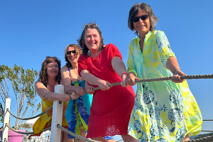 Vier Frauen in bunten Strandkleidern stehen auf einem Steg, halten gemeinsam ein Seil und ziehen spielerisch daran.