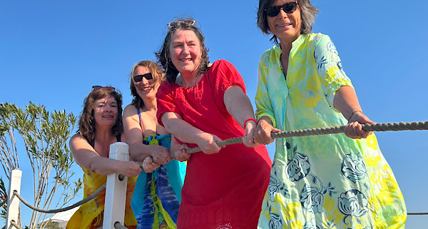 Vier Frauen in bunten Strandkleidern stehen auf einem Steg, halten gemeinsam ein Seil und ziehen spielerisch daran.