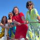 Vier Frauen in bunten Strandkleidern stehen auf einem Steg, halten gemeinsam ein Seil und ziehen spielerisch daran.