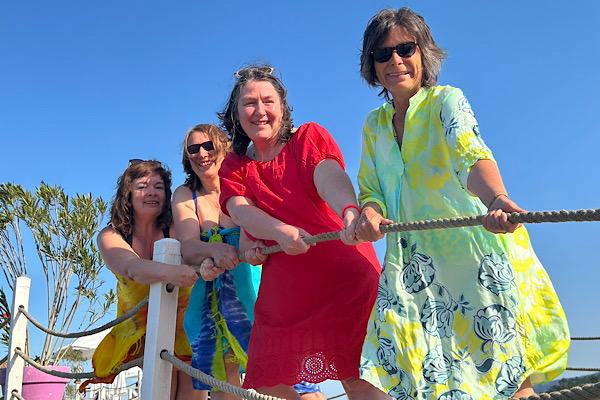 Vier Frauen in bunten Strandkleidern stehen auf einem Steg, halten gemeinsam ein Seil und ziehen spielerisch daran.