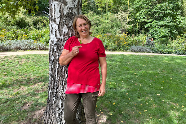 Anita steht lächelnd an einer Birke in der Natur und hält ein Glückskleeblatt in der Hand.