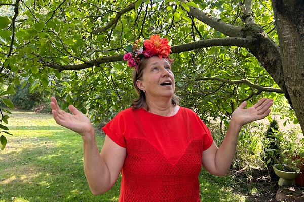 Anita Griebl mit Blumen im Haar, offene Arme zur Baumkrone, verbunden mit Natur und Herzenergie – Ausdruck ganzheitlicher Wahrnehmung.