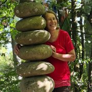 Anita Griebl steht im Nepalpark neben einer Steinsäule, blickt lächelnd hervor und strahlt Ruhe, Offenheit und Verbundenheit aus.