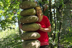 Was hält dich davon ab zu starten_ Anita Griebl steht im Nepalpark neben einer Steinsäule, blickt lächelnd hervor und strahlt Ruhe, Offenheit und Verbundenheit aus.