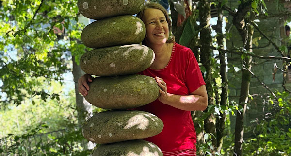 Anita Griebl steht im Nepalpark neben einer Steinsäule, blickt lächelnd hervor und strahlt Ruhe, Offenheit und Verbundenheit aus.