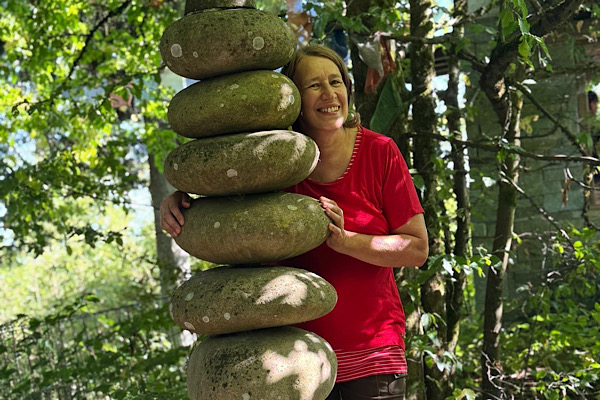 Anita Griebl steht im Nepalpark neben einer Steinsäule, blickt lächelnd hervor und strahlt Ruhe, Offenheit und Verbundenheit aus.