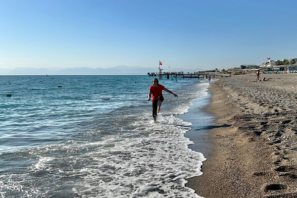 Frau geht am Strand durch flache Wellen, Meer und Horizont im Hintergrund.