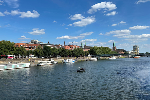 Blick von einer Brücke über die sonnige Weser auf die Stadt Bremen, mit klarer Sicht auf Ufer, Gebäude und Sommerhimmel.
