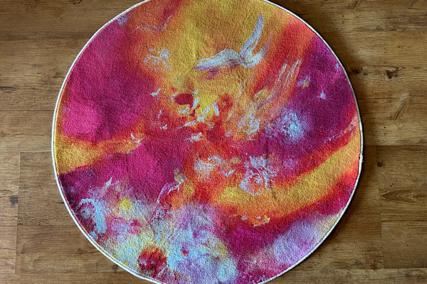 Runder, farbenfroher Teppich mit leuchtenden Rot‑, Orange‑, Gelb‑ und Weißtönen, die sich abstrakt vermischen und Bewegung ausdrücken.