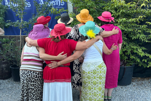 Monatsrückblick Juli 2026 Sieben Frauen stehen in zwei Reihen mit bunten Hüten nebeneinander und legen einander die Arme seitlich um, von hinten fotografiert.