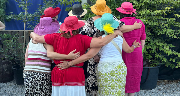 Sieben Frauen stehen in zwei Reihen mit bunten Hüten nebeneinander und legen einander die Arme seitlich um, von hinten fotografiert.