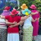 Sieben Frauen stehen in zwei Reihen mit bunten Hüten nebeneinander und legen einander die Arme seitlich um, von hinten fotografiert.