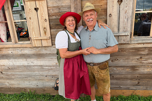 Anita und Karl stehen in Tracht vor einem Holzhaus; Anita trägt einen roten Hut, Karl einen Strohhut.