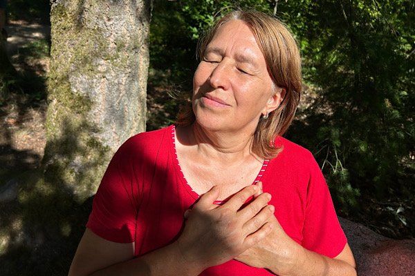 Anita Griebl sitzt mit geschlossenen Augen auf einer Steinbank, die Hände am Herzen, während die Sonne ihr Gesicht erhellt – ein Moment tiefer innerer Verbundenheit.