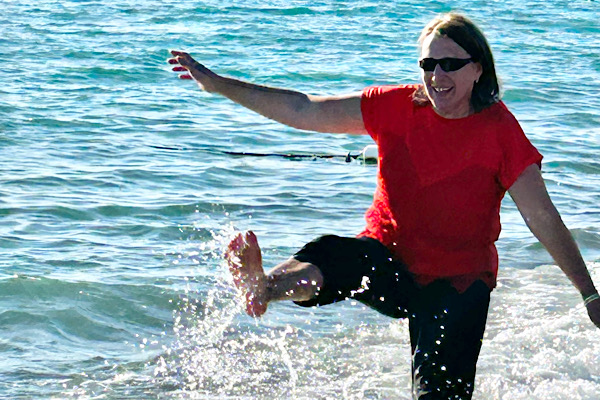 Anita Griebl steht im Meer und spritzt mit den Füßen Wasser hoch – ein Moment von Leichtigkeit, Freude und dem Schritt vom Wissen ins Tun.
