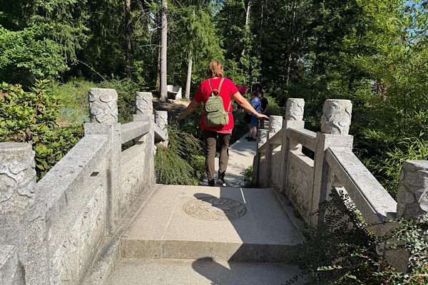 Anita mit Rucksack geht über eine Steinbrücke im Nepalpark; sie ist von hinten zu sehen, umgeben von Garten und Natur.