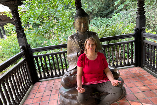 Anita sitzt vor einer Buddha‑Statue, hält die Hände in Meditationshaltung; die Statue steht unter einer Überdachung mit Geländer, im Hintergrund ist der Garten des Nepalparks zu sehen.