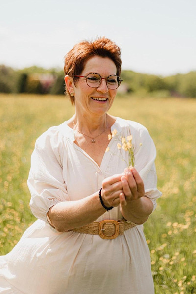 Karola steht in einem weißen Kleid auf einer Wiese und hält hellgelbe Blumen in den Händen; ruhige, naturverbundene Stimmung.