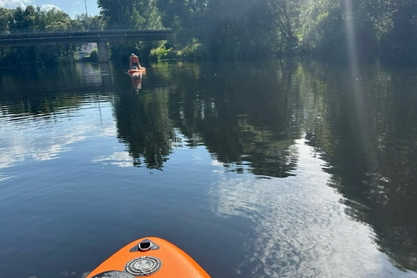 Ein ruhiger See mit der Spitze eines Paddelbootes im Vordergrund und einer Person auf einem Paddleboard im Hintergrund – ein Bild für Balance, Weite und innere Leichtigkeit.