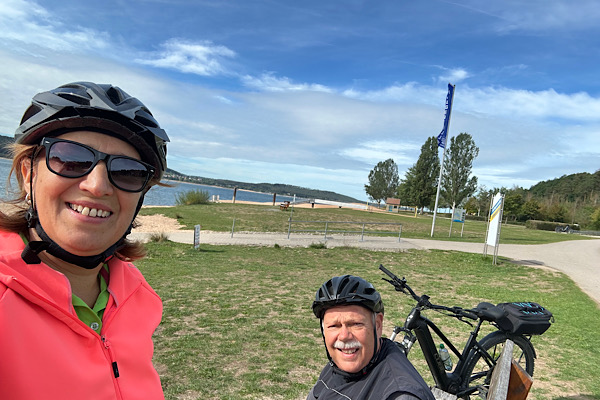 Selfie von zwei Personen mit Fahrradhelmen; im Hintergrund sind der Brombachsee und die Anlegestelle des Schiffs zu sehen.