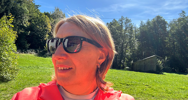 Anita Griebl mit Sonnenbrille schaut seitlich aus dem Bild; hinter ihr liegen eine Wiese, ein Wald und Sonnenstrahlen im Hintergrund.