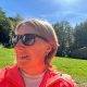 Anita Griebl mit Sonnenbrille schaut seitlich aus dem Bild; hinter ihr liegen eine Wiese, ein Wald und Sonnenstrahlen im Hintergrund.