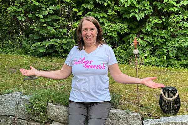 Anita Griebl sitzt auf einer Steinmauer im Garten und trägt ein weißes T‑Shirt mit der Aufschrift ‚Herzensmensch‘ – ein Moment von Offenheit und Einladung.