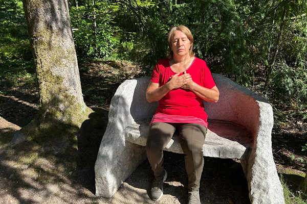 Anita Griebl sitzt mit geschlossenen Augen auf einer Steinbank, die Hände am Herzen – ein Moment bewusster Selbstfürsorge und innerer Ruhe.
