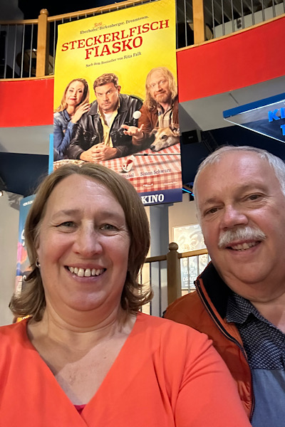 Selfie von zwei Personen vor dem Kinoplakat des Films Steckerlfischfiasko; darauf sind die drei Schauspieler Franz, Rudi und Susi zu sehen.