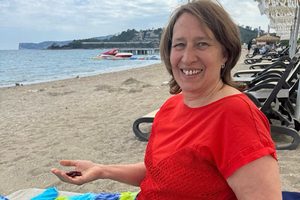 Anita Griebl am Strand auf einer Liege, lächelnd zur Kamera, mit Vitalstoff‑Kapseln in den Händen – ein Moment von Leichtigkeit, Pflanzenkraft und innerer Klarheit.