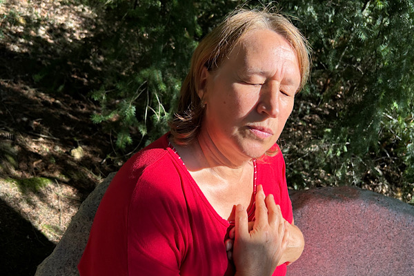 Anita mit geschlossenen Augen sitzt im Sonnenlicht, die Hände auf dem Herzen, ein Moment innerer Verbundenheit.