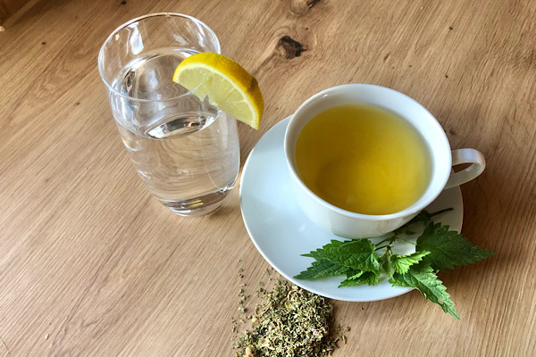 Glas Wasser mit Zitronenscheibe und eine Tasse Brennnesseltee auf einem Teller – natürliche Unterstützung für sanfte Körperreinigung.